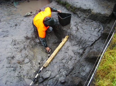 Harri Blomberg tar upp vatten med en skopa på 1,55 meters djup, för att tillsammans med arkeologer inom Kungahällaprojektet kunna dokumentera en bronsålderslämning i Ytterby, Bohuslän.